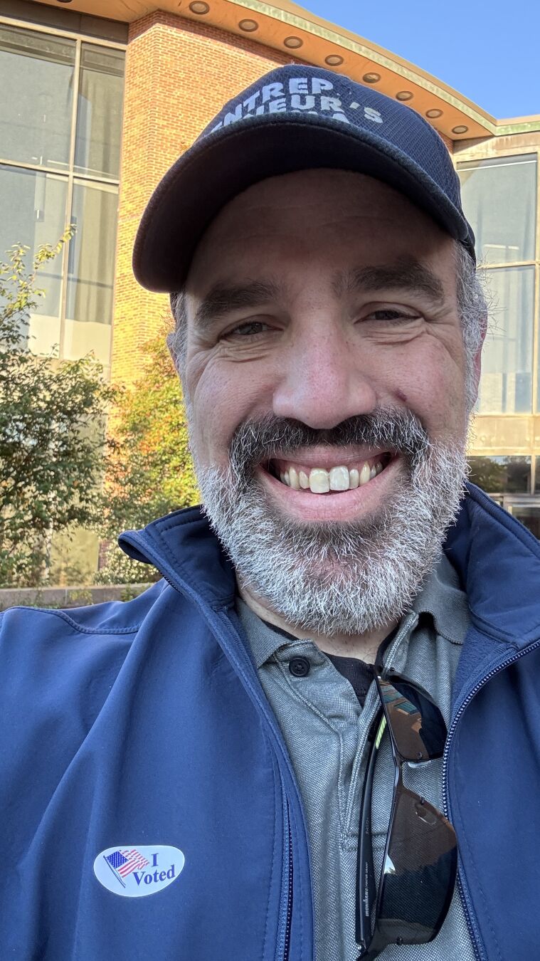 Me wearing a I voted sticker on my navy blue jacket with a gray polo underneath.

I'm wearing an Entrepreneur's Enigma podcast hat.

Gray bearded and smiling!